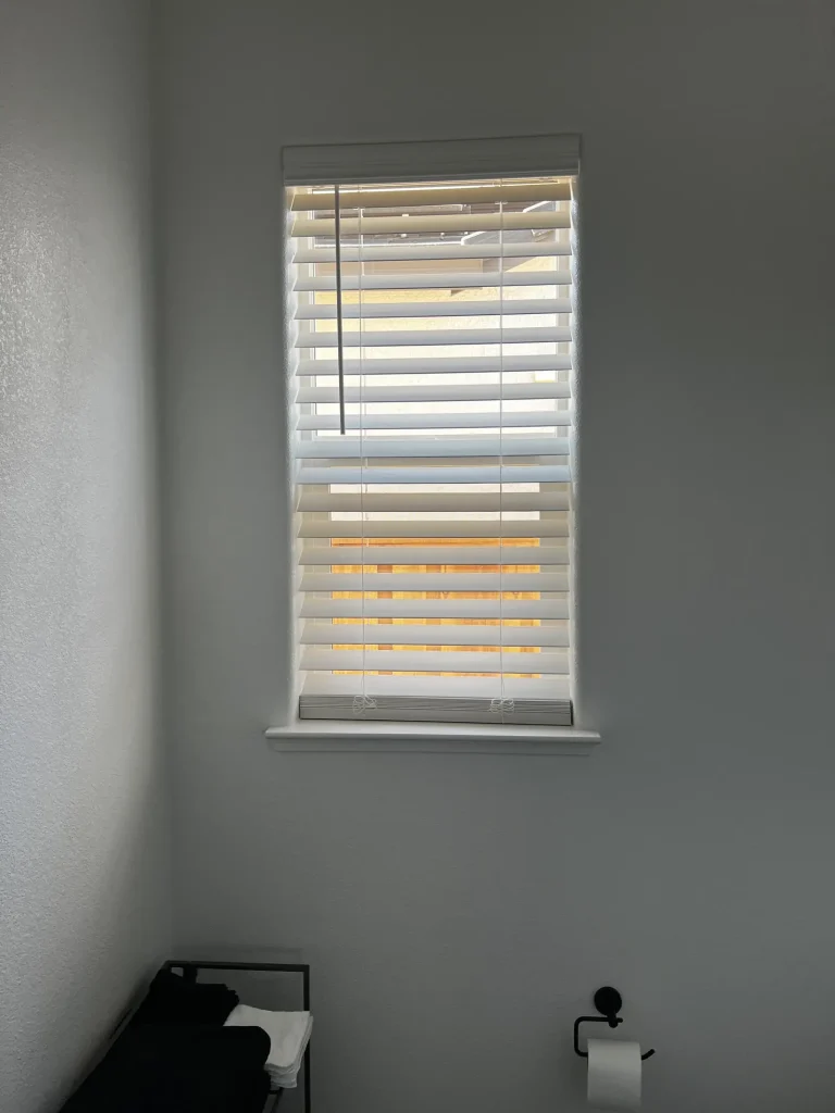 White faux wood blinds fitted inside a tall bathroom window in Lathrop, slats tilted open and the valance closing off the headrail