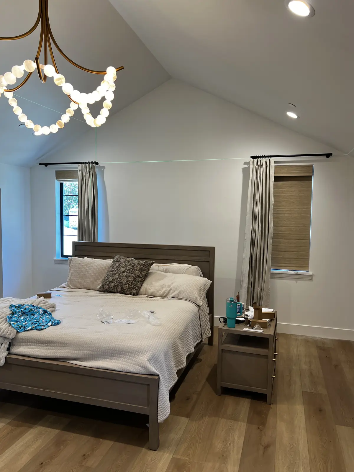 Linen drapery panels and woven wood shades hung at two bedroom windows in Lodi, under a vaulted ceiling with the laser line still lit