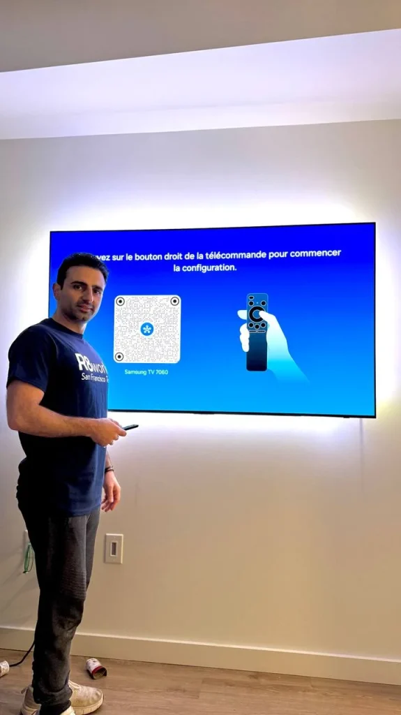 Flat screen TV mounted above a wooden floor in a Sacramento home, displaying a blue setup screen as the technician tests the remote.